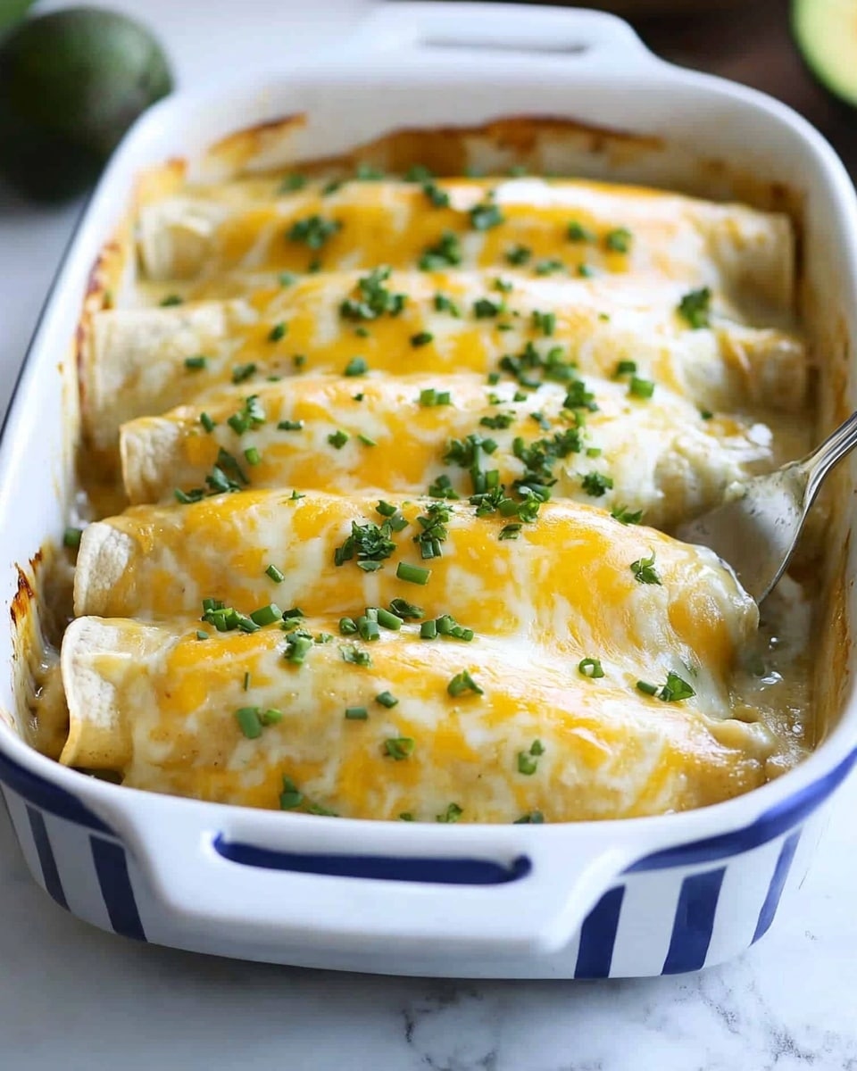 Smothered Green Chile Chicken Burritos, green chile chicken burritos, cheesy chicken burritos, easy Mexican dinner recipes, flavorful burritos The dish shows a white baking dish with blue stripes on the outside, filled with five rolled enchiladas covered in melted cheese. Each enchilada has a soft, light yellow tortilla base, covered fully with thick, creamy, white sauce and topped with a golden-yellow melted cheese layer that stretches slightly as one enchilada is lifted by a spatula. Small green chopped chives are sprinkled over the cheese, adding a fresh color contrast. The background is a white marbled surface with blurred avocado halves and a small bowl visible. The