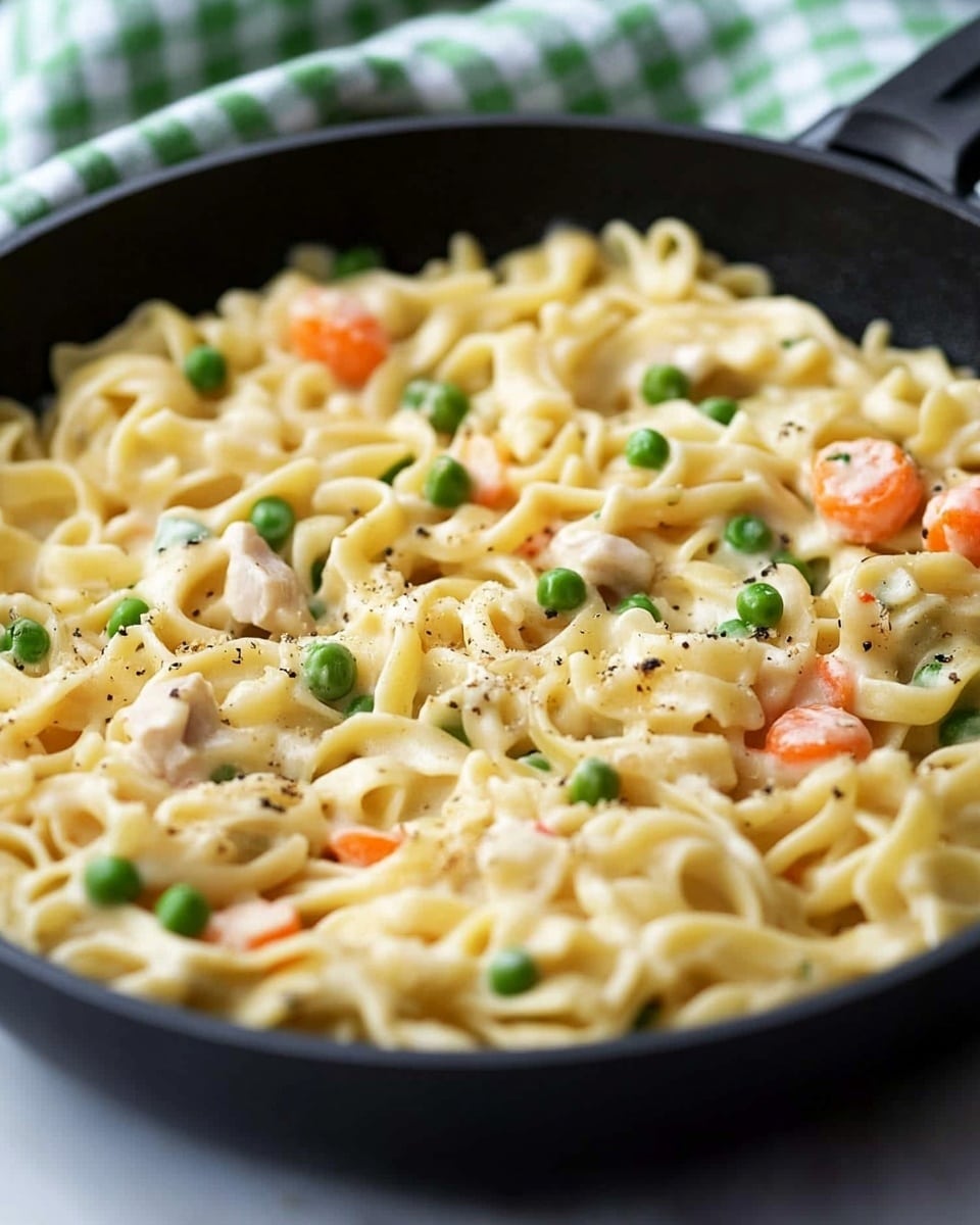 A close-up view of a black pan filled with creamy pasta. The pasta is light yellow with a soft and smooth texture, mixed with small pieces of orange carrots and bright green peas scattered evenly throughout. There are also light pieces of tender white meat interspersed in the mix. The pan sits on a white marbled surface, and a green and white checkered kitchen towel is partially wrapped around the pan s handle. The sauce looks thick and coats the pasta well, giving a rich appearance. Photo taken with an iphone --ar 4:5 --v 6.1