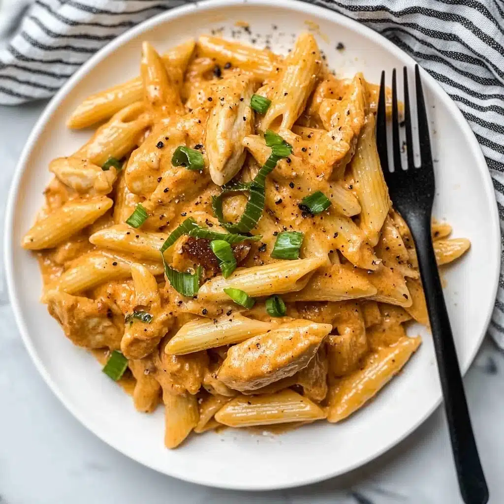 One Pot Creamy Cajun Chicken Pasta, Cajun Chicken Pasta, Creamy Cajun Pasta, Easy Cajun Chicken Dinner, One Pot Pasta Recipes A white round plate holds a creamy pasta dish made of several layers: at the bottom are pieces of light brown cooked chicken and a soft, light orange sauce mixed with whole and chopped penne pasta which is pale yellow. Scattered on top are bright green sliced scallions adding freshness, and small black pepper flakes are sprinkled around. A black fork rests on the right side of the plate touching the pasta, and the whole plate is set on a white marbled surface with a black and white striped cloth nearby.