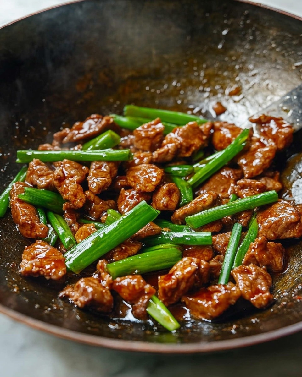 Mongolian Chicken, Mongolian Chicken recipe, Asian chicken dishes, quick weeknight dinner, savory chicken recipes Inside a dark metal wok on a white marbled surface, small pieces of browned meat are mixed with bright green scallions. The meat looks shiny and cooked in a thick brown sauce that pools slightly at the bottom. The scallions are cut into long thin pieces and scattered evenly on top, adding a fresh contrast in color and texture. The wok shows signs of use, with some light marks and a textured surface inside, and a metal spatula edge is visible at the top right corner.