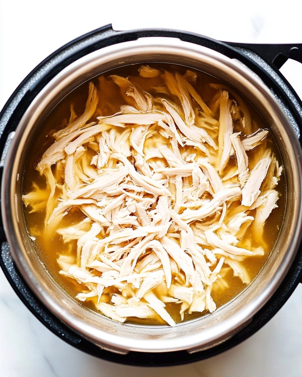 Instant Pot Shredded Chicken, easy shredded chicken, quick chicken recipes, pressure cooker chicken, versatile chicken dishes The image shows a close-up top view of shredded light beige chicken resting in a shallow layer of golden-brown broth inside a shiny silver metal pot. The chicken pieces are long, thin, and stringy, scattered unevenly throughout the pot. The pot has a black outer rim and handles, and it sits on a bright white marbled surface. The light reflects softly off the broth and the moist chicken, creating a fresh and clean look.