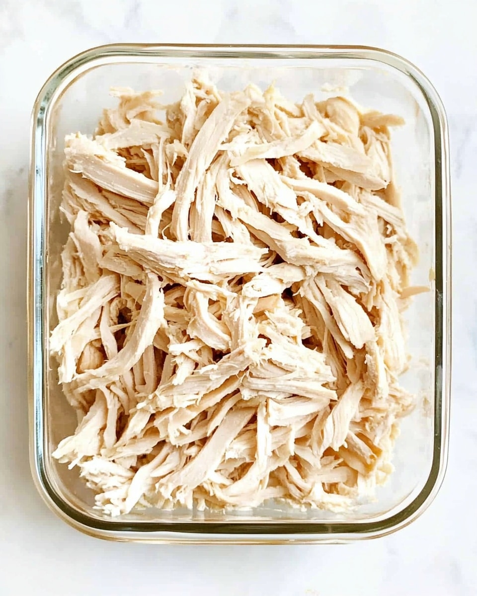 Instant Pot Shredded Chicken, easy shredded chicken, quick chicken recipes, pressure cooker chicken, versatile chicken dishes The image shows a clear glass rectangular container filled with shredded cooked chicken. The shredded chicken is light beige, with soft, fibrous texture pieces layered loosely to fill the container evenly from top to bottom. The chicken strands are thin and varied in size and shape, creating a natural, slightly uneven surface. The container sits on a white marbled surface.