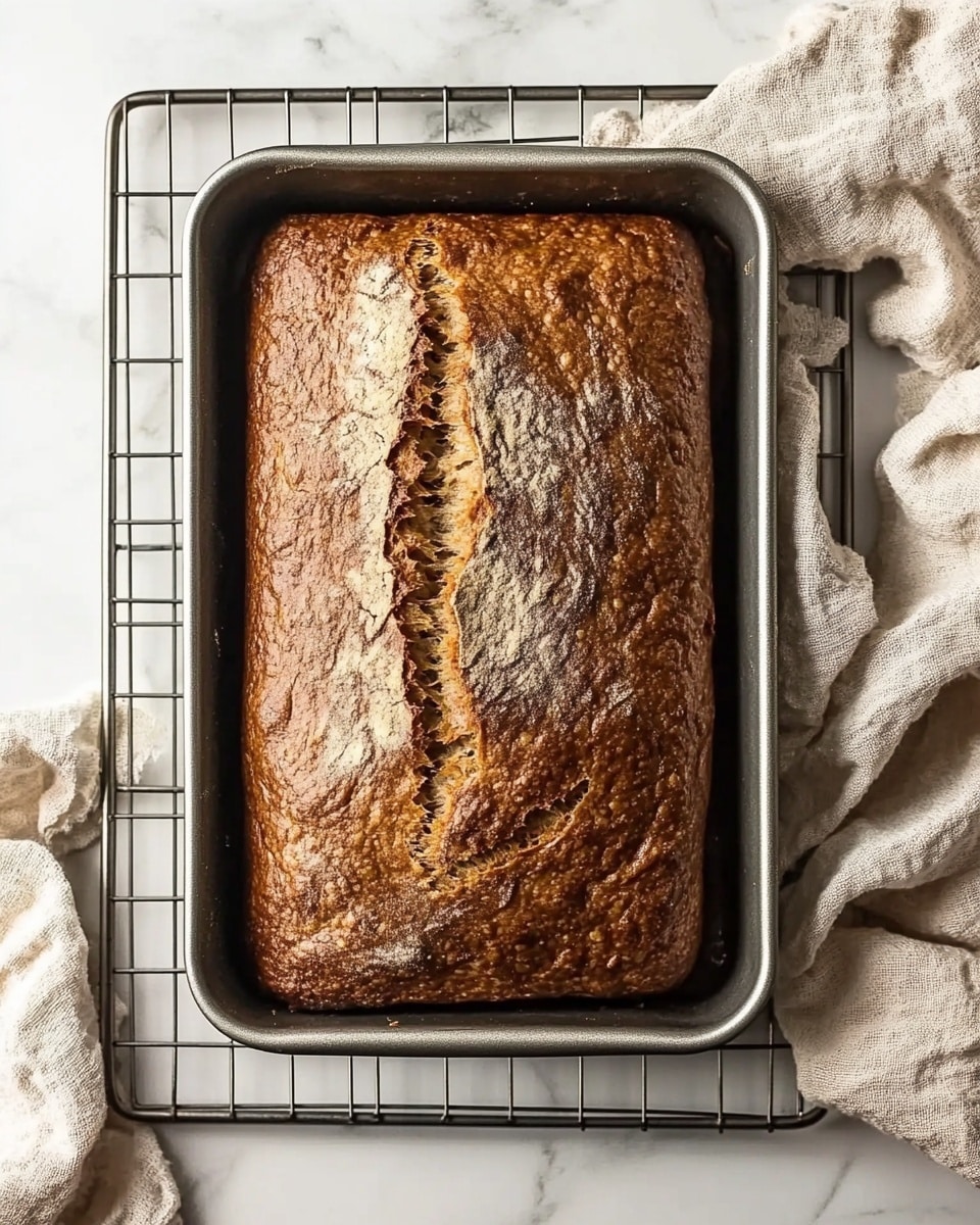 Simple Homemade Banana Bread, easy banana bread recipe, quick banana bread, moist banana bread, beginner banana bread A rectangular loaf of brown bread with a cracked top surface rests in a silver metal loaf pan, placed on a metal cooling rack over a white marbled texture. The bread's crust is evenly baked with a golden brown color and some darker spots, showing a rough texture. Next to the pan, a beige fabric cloth is casually placed, adding a soft touch to the scene.