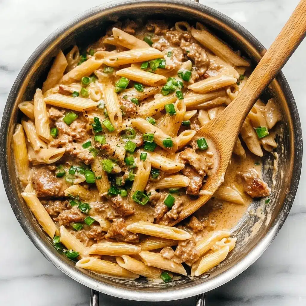 One Pot Creamy Cajun Chicken Pasta, Cajun Chicken Pasta, Creamy Cajun Pasta, Easy Cajun Chicken Dinner, One Pot Pasta Recipes A stainless steel pan filled with cooked penne pasta mixed with small pieces of browned cooked meat and chopped green onions scattered on top. The pasta and meat are coated in a creamy light brown sauce that looks smooth and rich. A wooden spoon rests inside the pan on the right side, partially covered with the pasta mixture. The pan is placed on a white marbled surface.