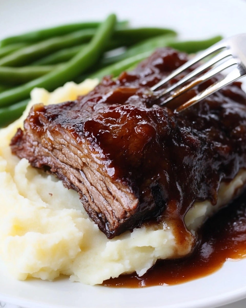 Instant Pot Beef Ribs with Broiled Crispy Bark, easy beef ribs recipe, quick pressure cooker beef ribs, tender beef ribs with crispy bark, flavor-packed beef ribs dinner The image shows a white plate with three food layers and a utensil in front. The bottom layer is mashed potatoes, which look smooth and creamy with a soft, fluffy texture, colored white to light yellow. On top of the mashed potatoes is a thick, dark brown barbecued meat piece with a shiny, caramelized sauce glaze and a slightly rough texture. To the right of the plate, green beans add a fresh and crisp green layer with a smooth surface. The background features a white marbled table surface and a blurred kitchen appliance, creating a cozy kitchen feel.