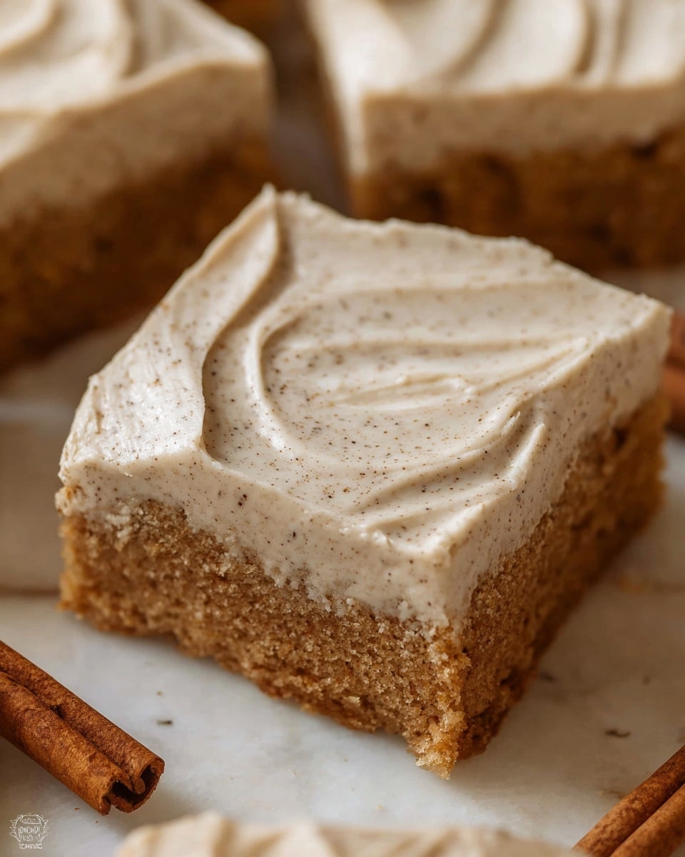 The image shows a close-up of two square pieces of moist brown cake with a thick layer of creamy light beige frosting on top. The frosting looks smooth and slightly swirled with a dusting of fine brown powder, likely cinnamon, over it. Each cake piece has one visible layer of cake and one layer of frosting. The cake texture appears soft and slightly crumbly. The setting is on a white marbled surface, with a blurry glass of dark coffee in the background and a fork with some cake crumbs on the side. Photo taken with an iphone --ar 4:5 --v 7